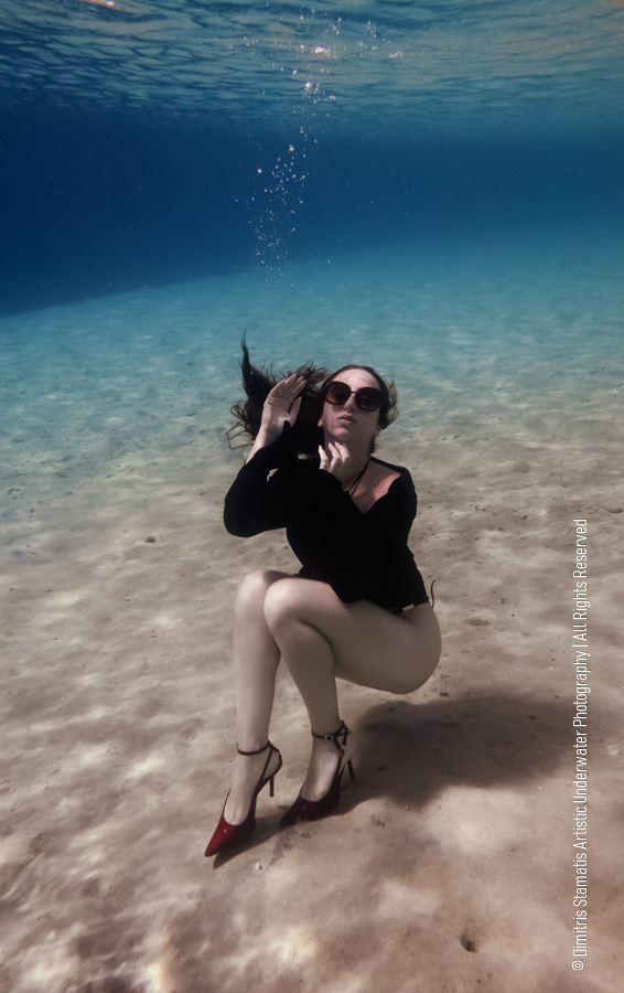 A girl underwater wearing a black top, red high‑heels, and sunglasses crouches on the sandy seabed.