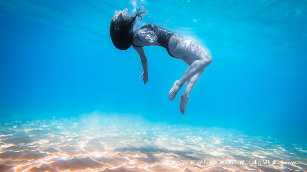 A young girl floats underwater above a sandy seabed, with hair drifting and sunlight filtering through the surface to create shimmering patterns on the sand and body.