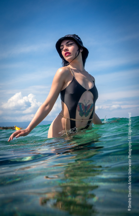A young girl in a black cut‑out swimsuit and bucket hat stands in the sea with the lower body submerged, a butterfly tattoo visible on the torso, under a bright blue sky with scattered clouds.