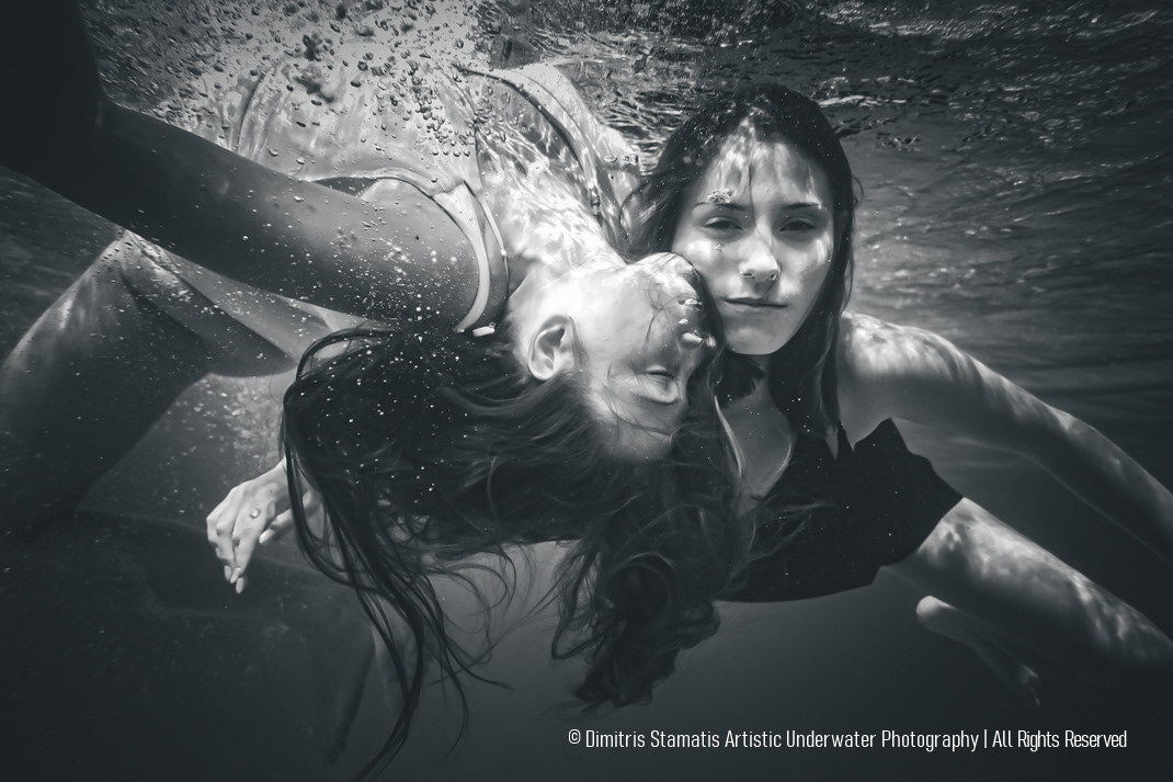 Two young girls submerged underwater, one facing the camera and the other turned sideways, with hair floating freely and bubbles surrounding them as light highlights their faces and bodies.