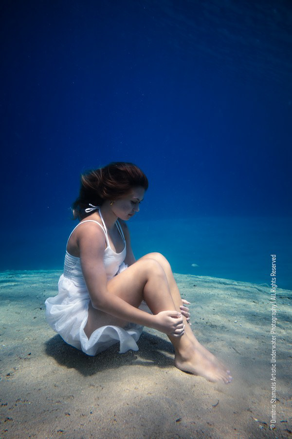 A girl sitting on the sandy seafloor underwater in a white dress, holding her legs.