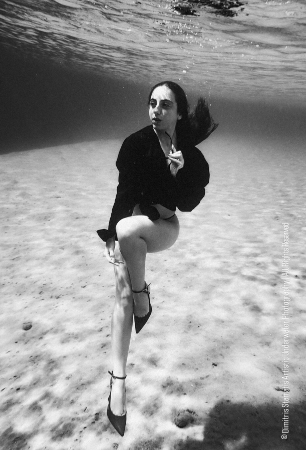 A girl underwater in a dark shirt and heels posing on the sandy seafloor as hair drifts.