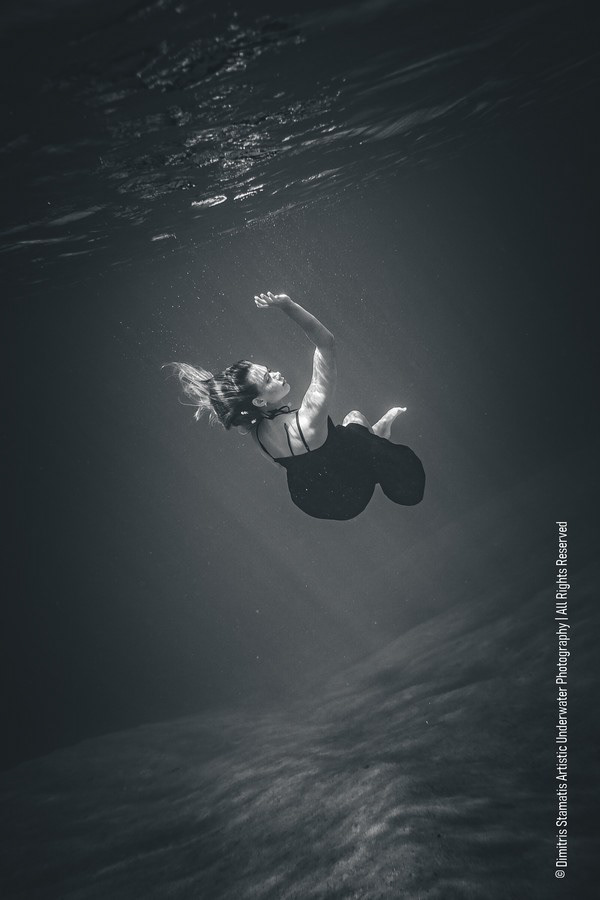 A woman wearing a dark outfit floats upward underwater with her arm extended as light rays filter through the surface and illuminate the sandy sea floor below.