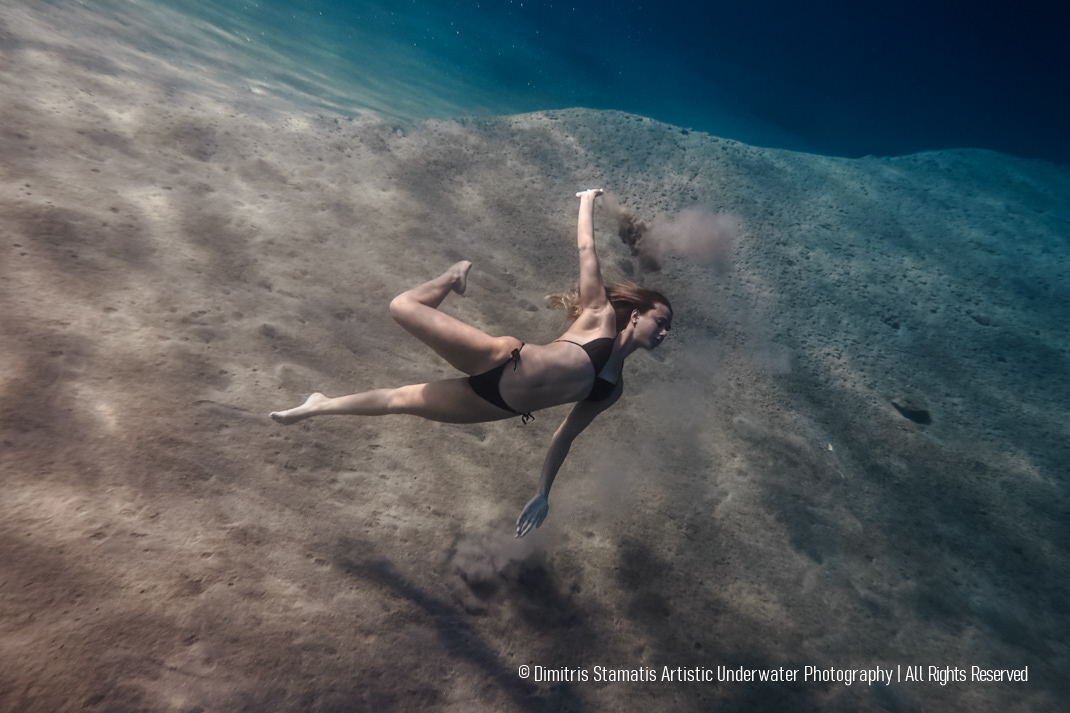A swimmer wearing a black swimsuit glides underwater close to the sandy seabed, creating a soft cloud of sand particles that rise through the clear blue water.