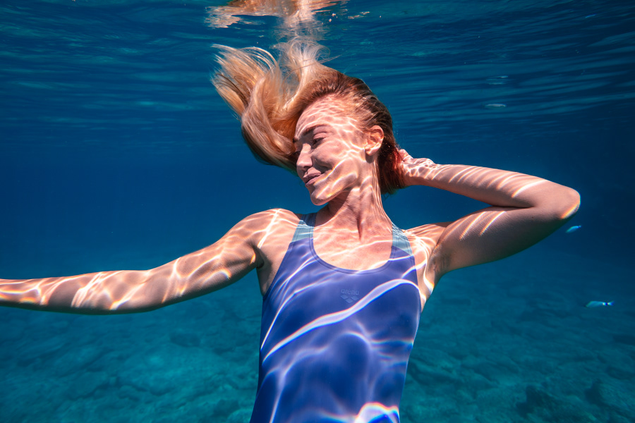 Şahika Ercümen underwater in a blue-lit scene with flowing hair and sunlit patterns on her skin.