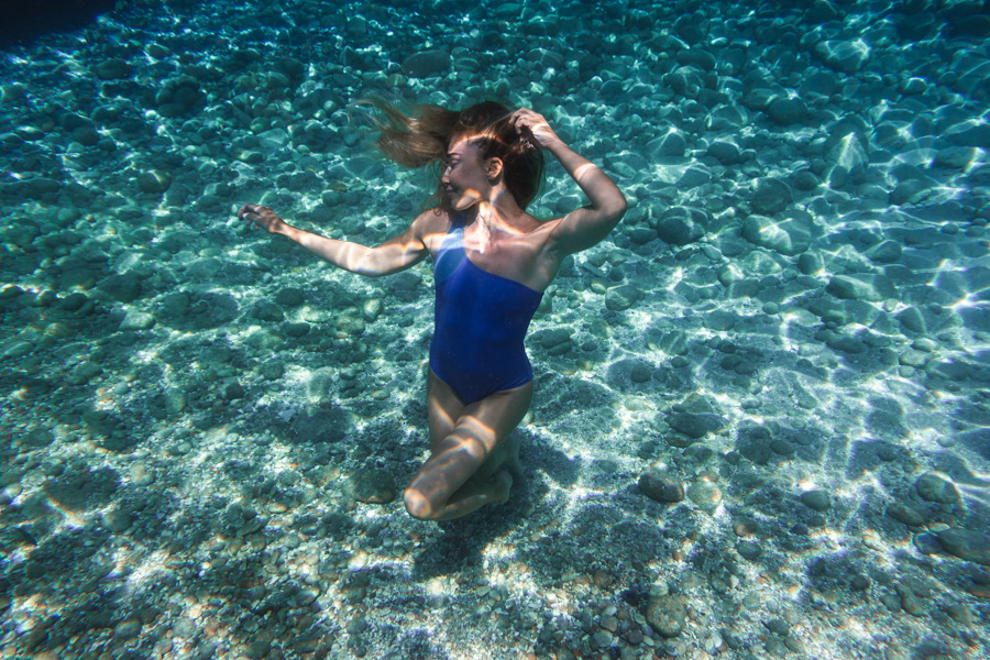 Şahika Ercümen posing underwater above a rocky shallow seabed with sunlit patterns across her skin.