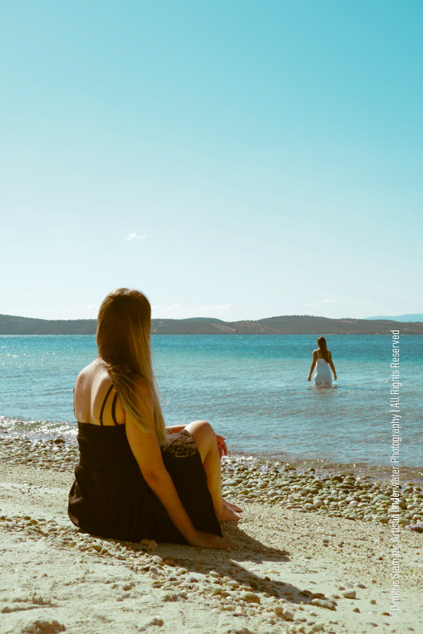 Two women at the seaside, one sitting on the sandy shore in a dark dress and the other standing in the shallow water in a light dress, looking toward the distant hills beneath a clear sky.