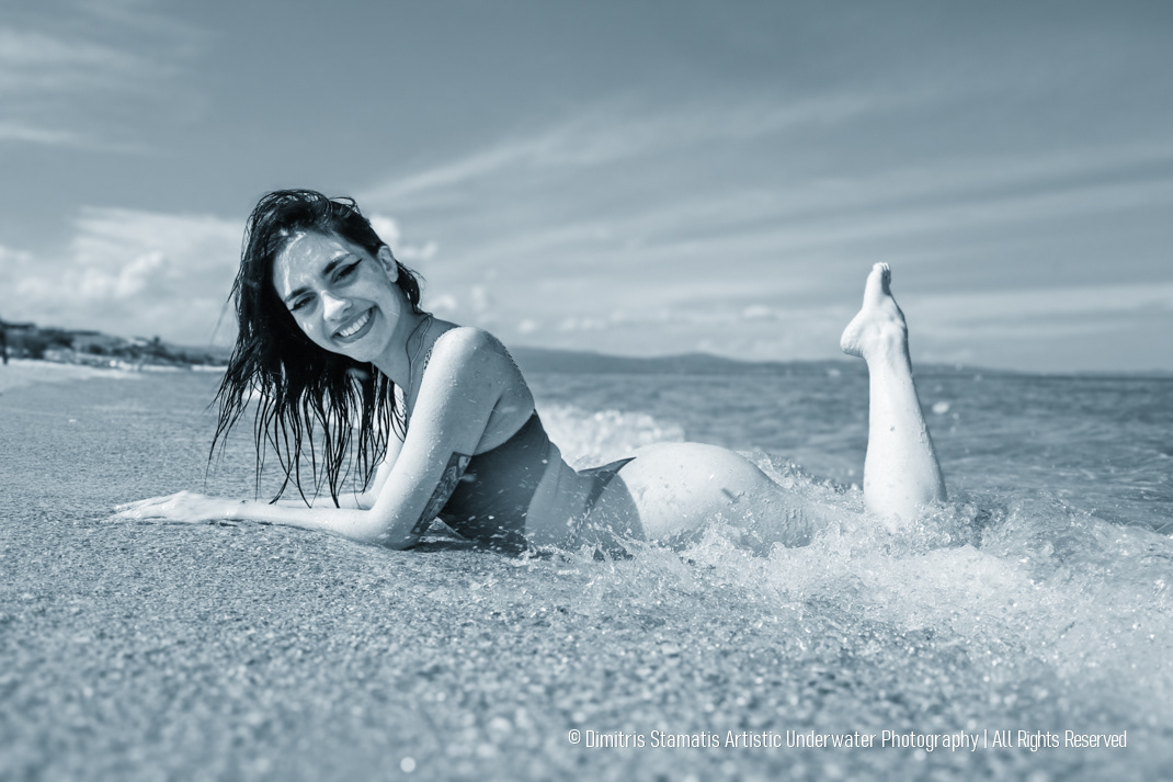 A young girl in a swimsuit lies at the water’s edge as waves wash over the sand, with the sea and distant coastline visible under a cloudy sky.