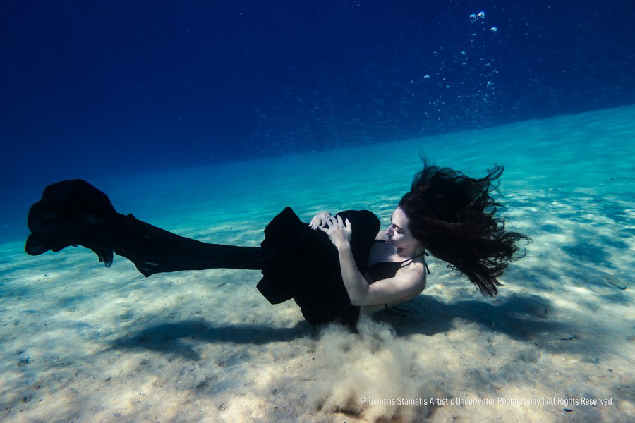 A girl underwater in a long black garment curled slightly above the sandy seafloor. 