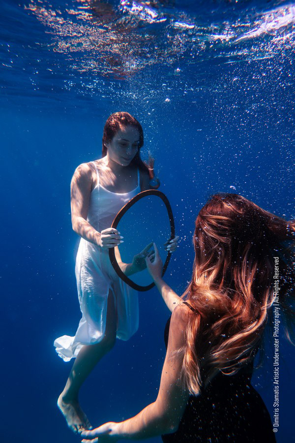 Two girls underwater reaching toward a circular mirror, one touching on its surface.
