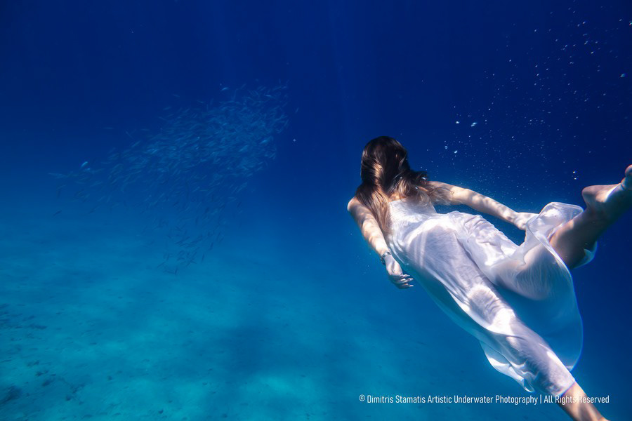 A young girl swimming underwater in a light blue dress, towards a school of fish.