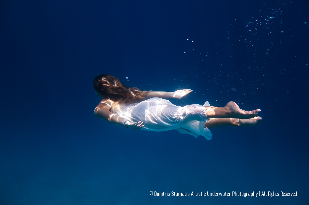 A young girl swimming underwater in a flowing white dress with long hair drifting gently in clear blue light.