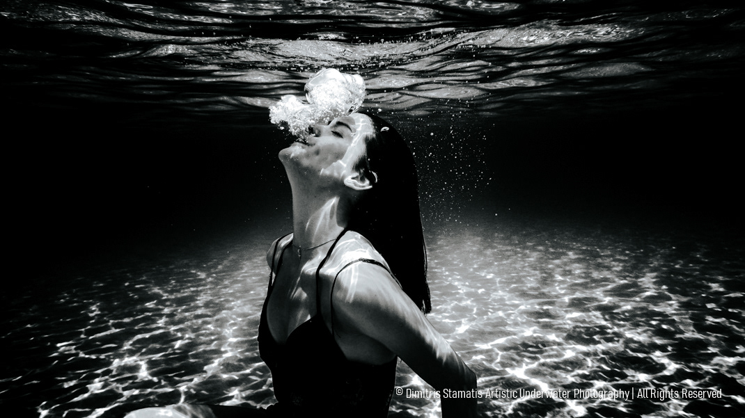 A young girl underwater with head tilted back as bubbles rise from her mouth and rippled light patterns move across the sandy seabed.