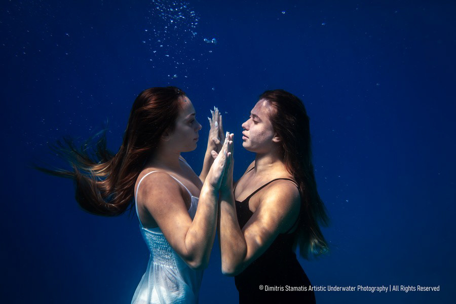 Two young girls underwater wearing black and white outfits touching hands in a deep blue sea.