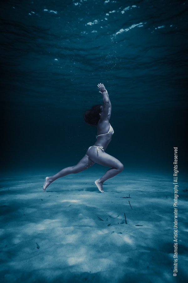 A young woman in a light bikini standing underwater in a yoga‑like pose above the sandy seabed as soft blue light filters through the water.