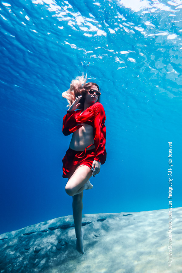 A young girl in a bright red outfit floating underwater just above the sandy seabed as sunlight filters through the clear blue water.