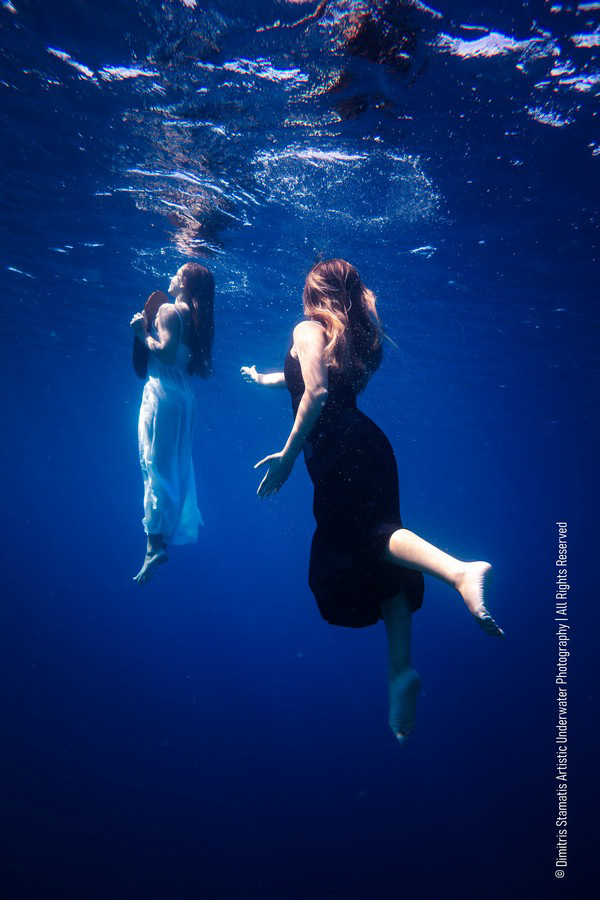 Two girls underwater in flowing black and white dresses drifting just below the surface.
