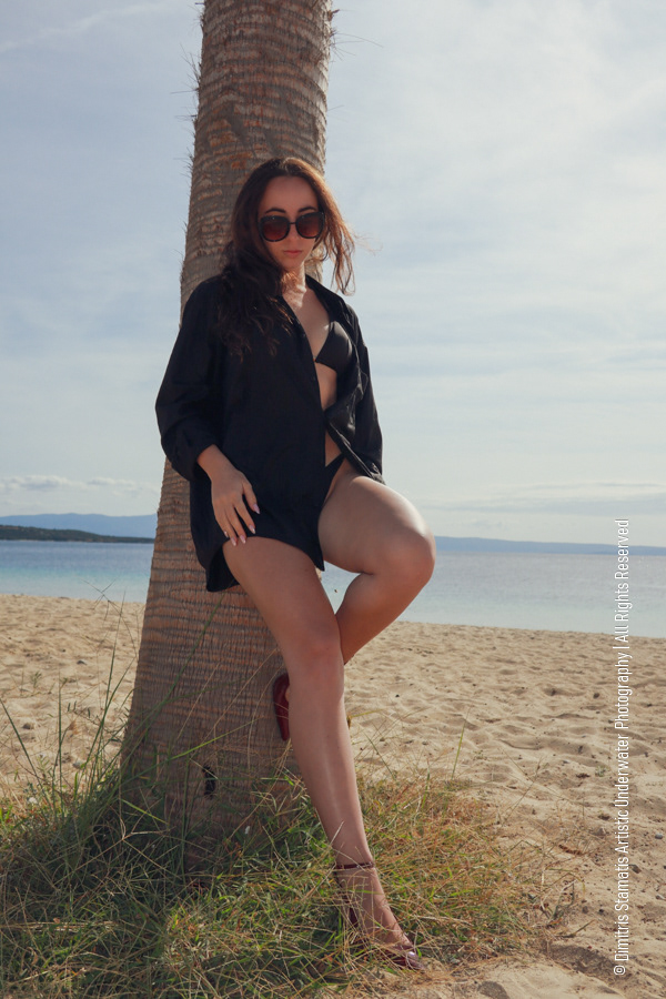 A girl on a sandy beach leaning on a palm tree wearing sunglasses, a dark shirt, and red heels.