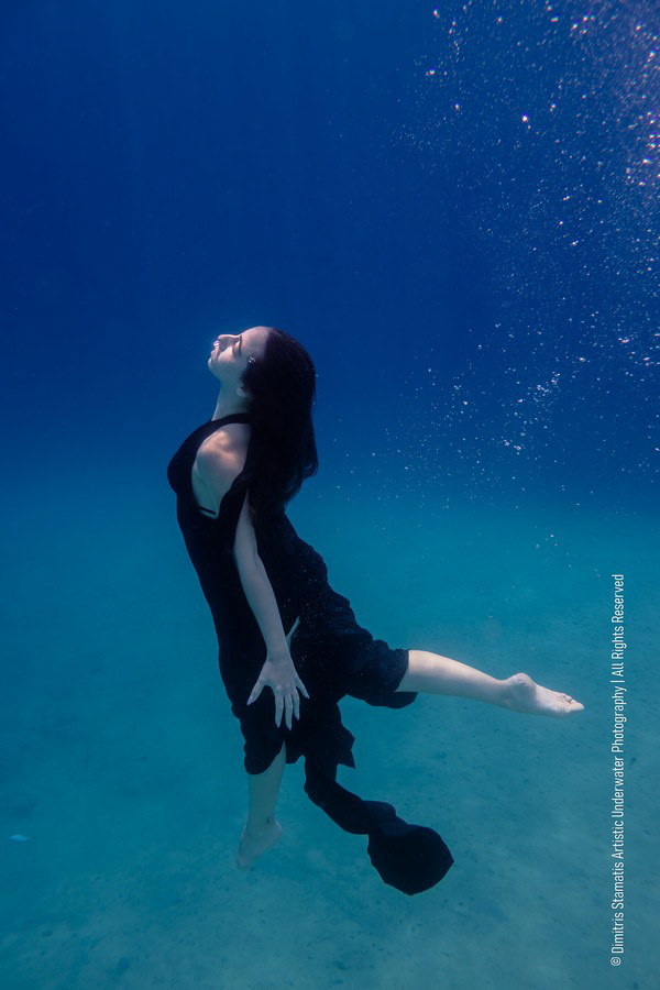 A girl underwater in a flowing black outfit floating above a sandy seabed as bubbles rise.