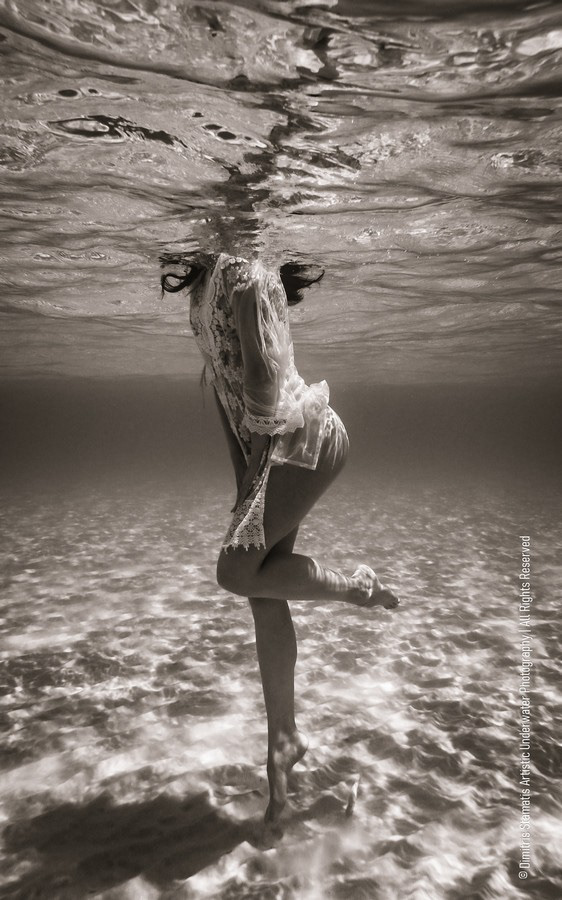 A woman wearing a light lace-trimmed dress stands on the sandy sea floor with her head submerged just below the water’s surface as sunlight patterns reflect around her.