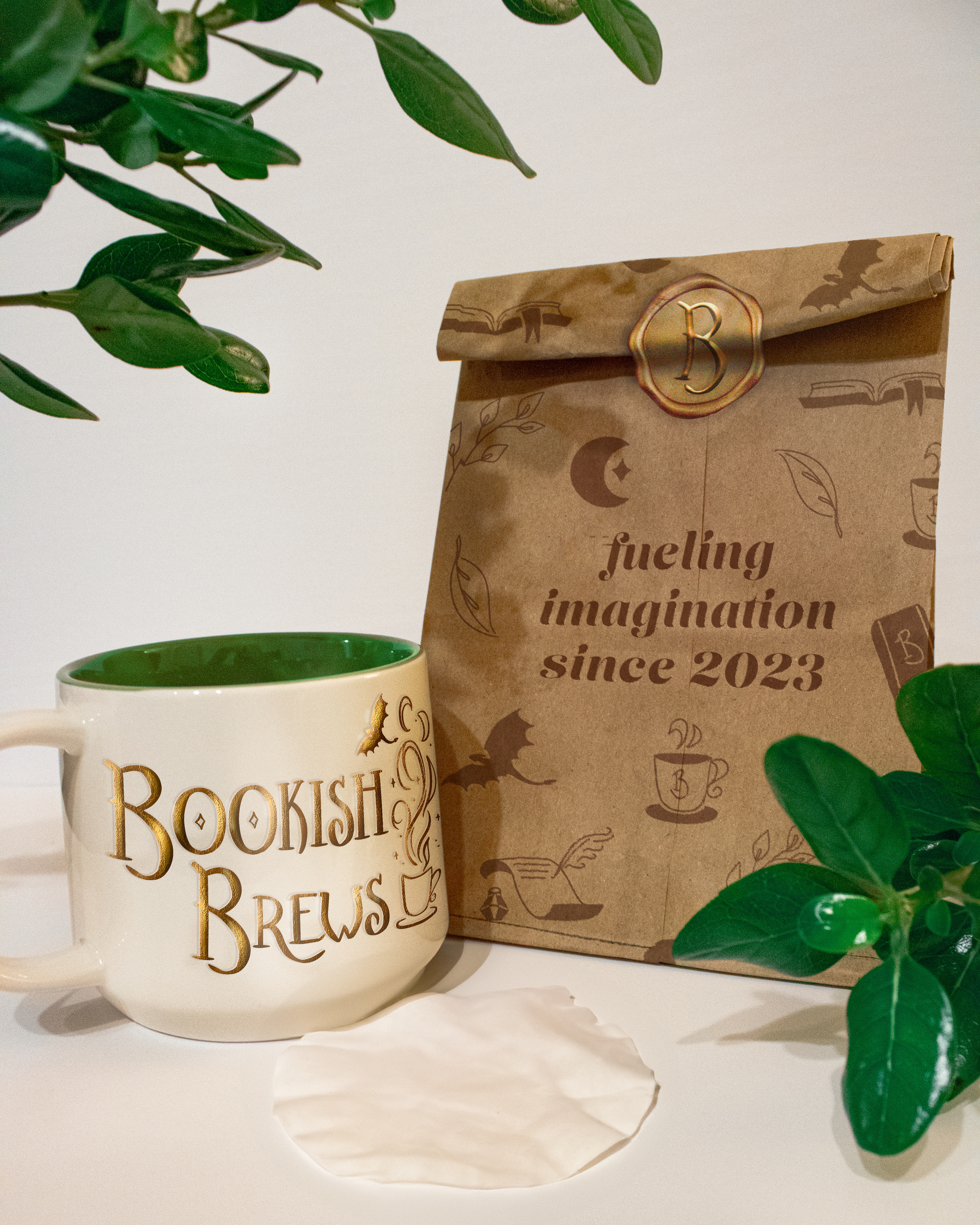 White mug with golden "Bookish Brews" logo next to a brown bag with decorative elements and the words "fueling imagination since 2023" on it.