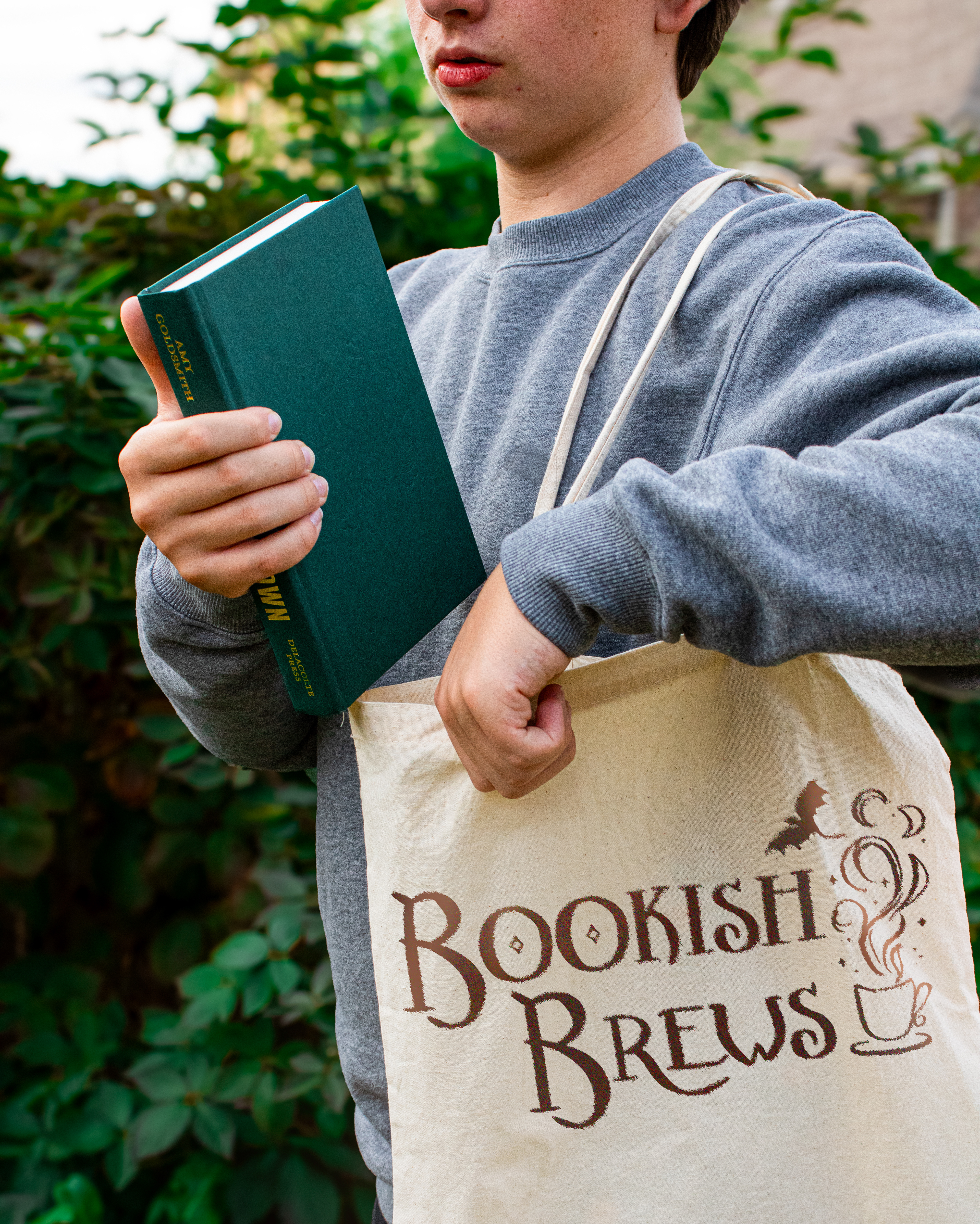 Model wearing a grey sweatshirt and holding a tan tote bag with the "Bookish Brews" logo.