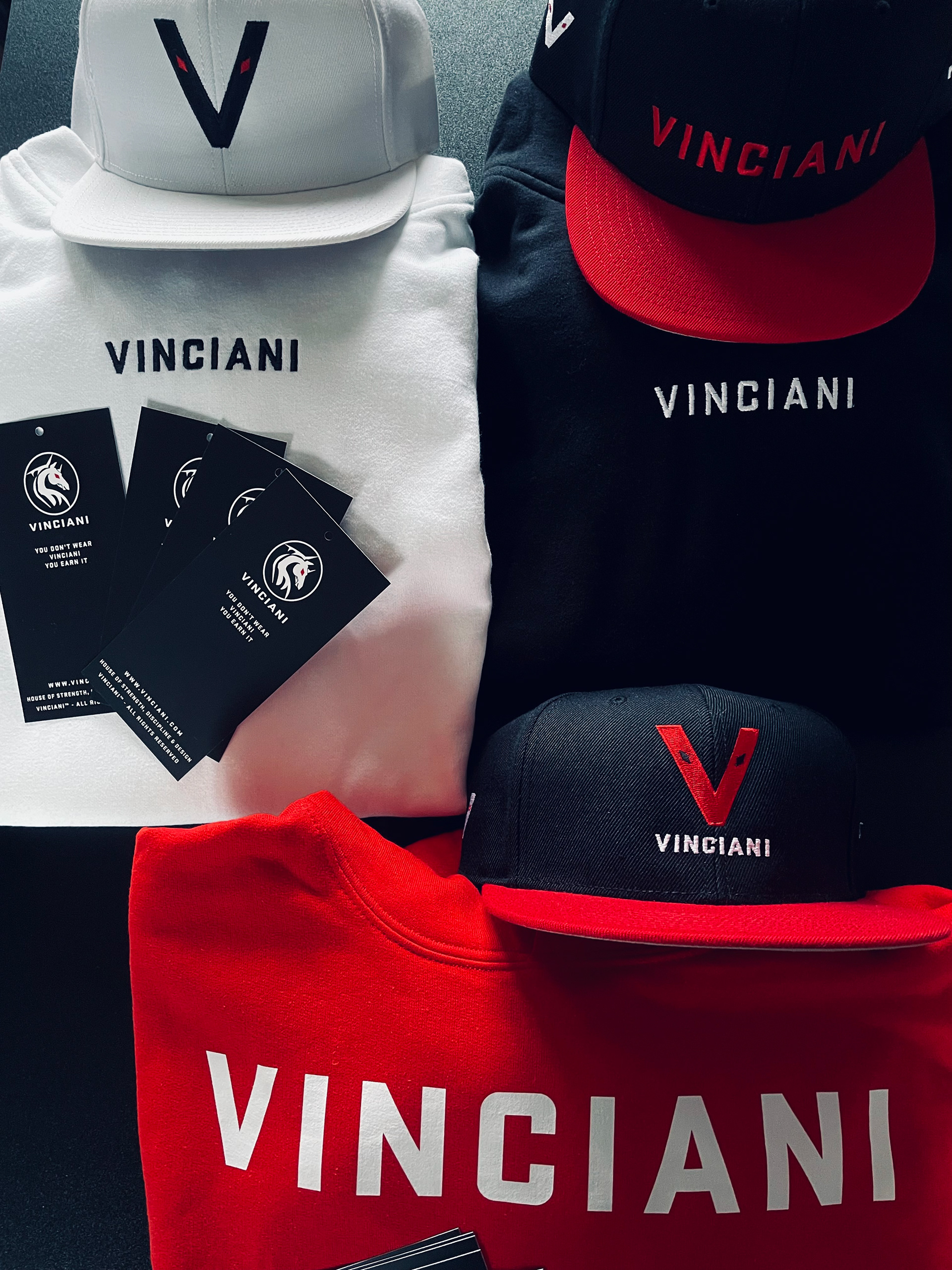 An overhead shot of a physical Vinciani collection featuring black, white, and red hoodies and caps alongside brand hashtags that carry the "You Don't Wear Vinciani, You Eran It" motto.