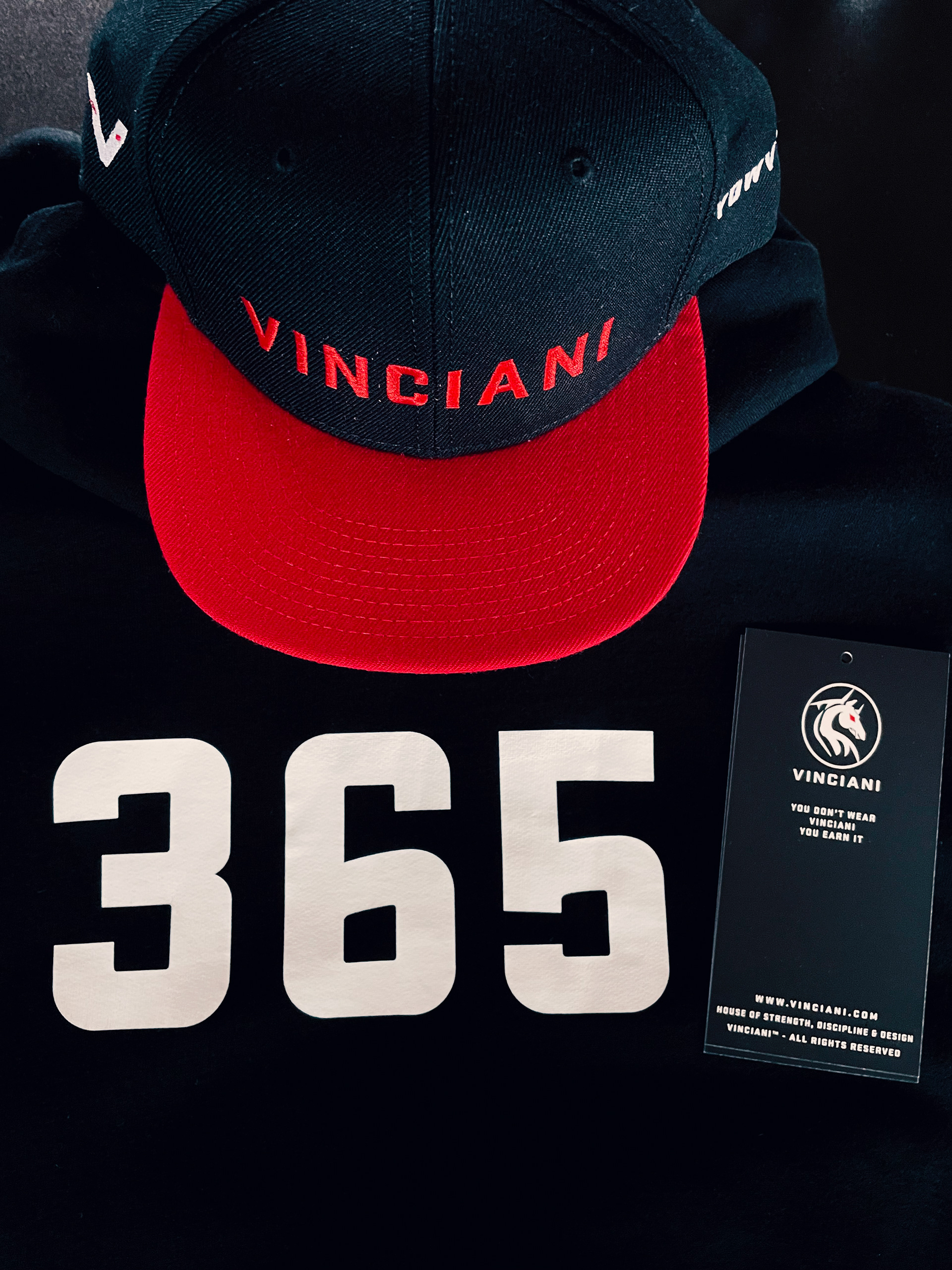 Flat lay of a black Vinciani hoodie jacket featuring the bold white 365 printed at the back, paired with a black and red embroidered Vinciani cap and branded tag.