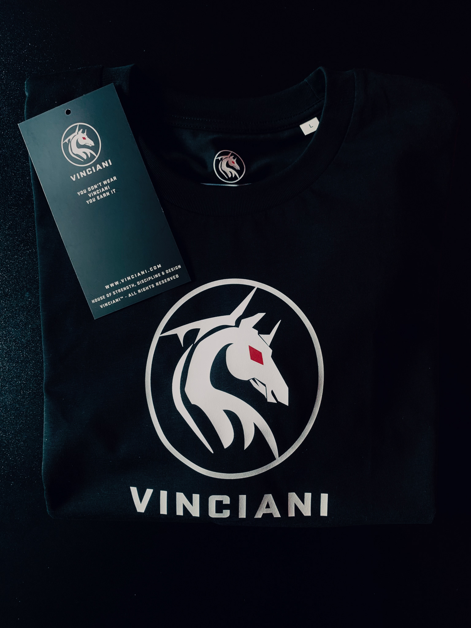 Black Vinciani t-shirt featuring the white horse emblem with red diamond eye, bold Vinciani branding, and signature hang tag with "You Don't Wear Vinciani, You Earn It"