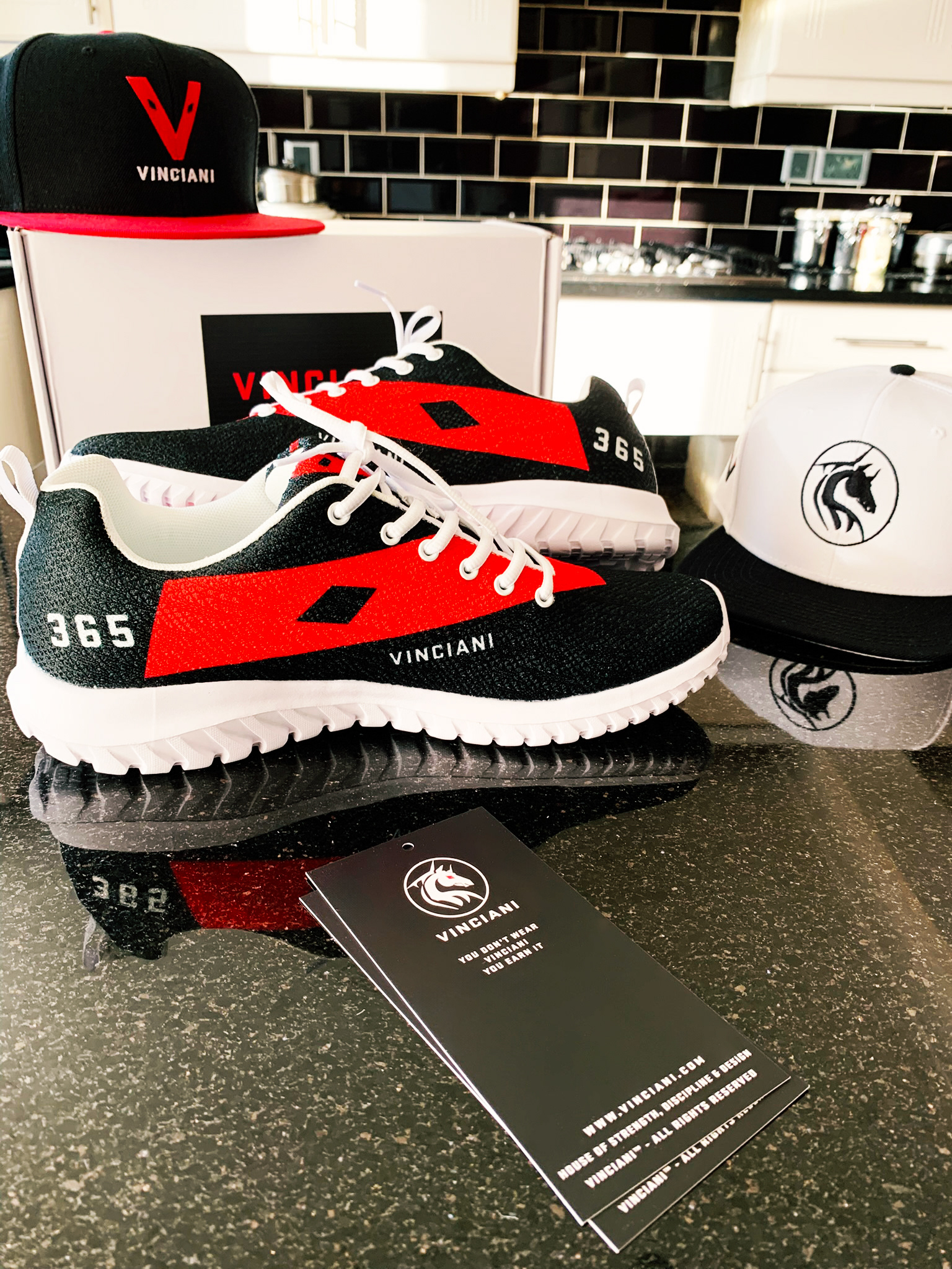A physical showcase of the Vinciani collection featuring the Vinciani Axion V Asymmetry sneakers, black and white branded caps, and a professional brand hangtag displayed on a dark surface.