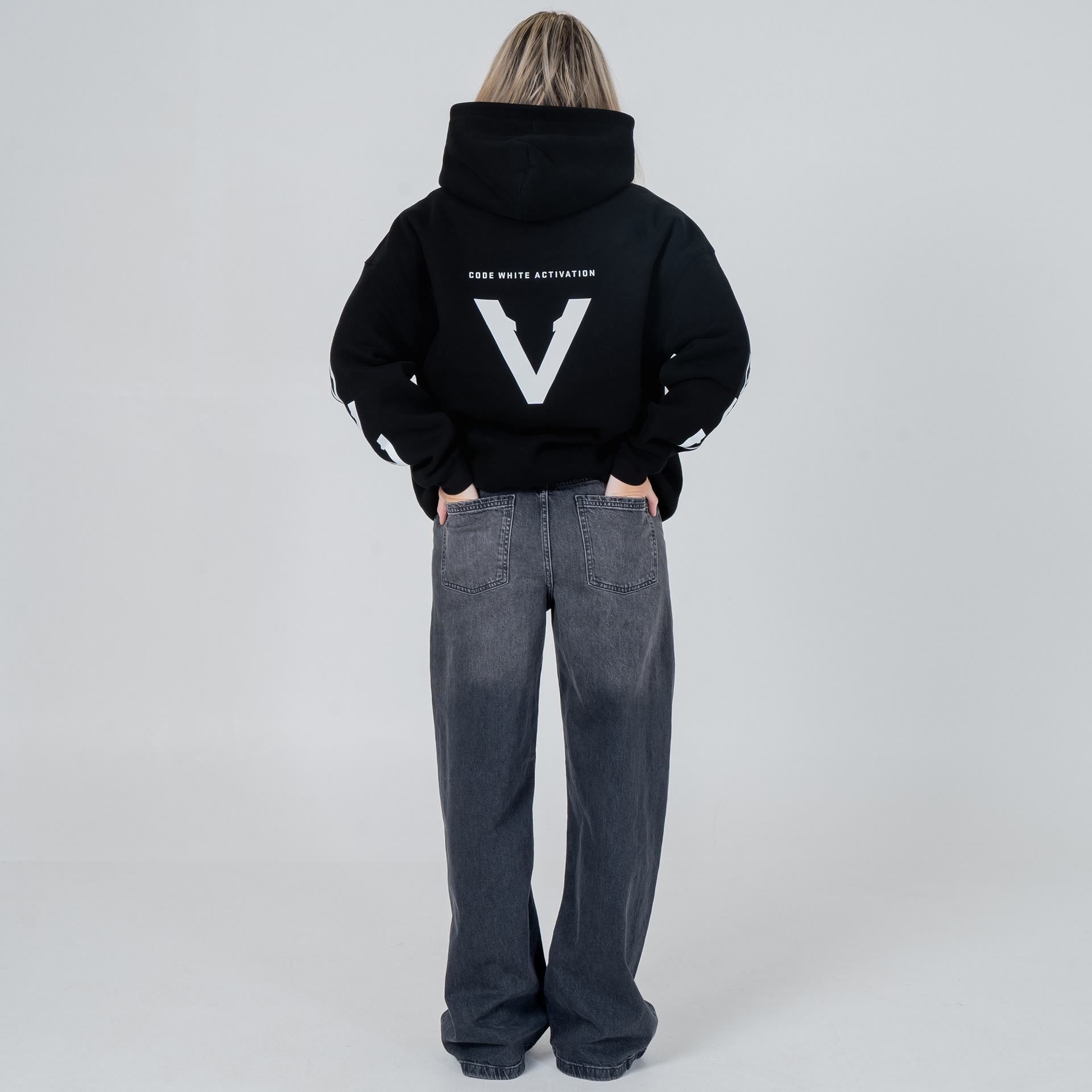 Rear view of female model wearing a black Vinciani Code White Activation hoodie featuring the large white V-Mark back graphic and sleeve graphics, styled with dark grey baggy jeans.