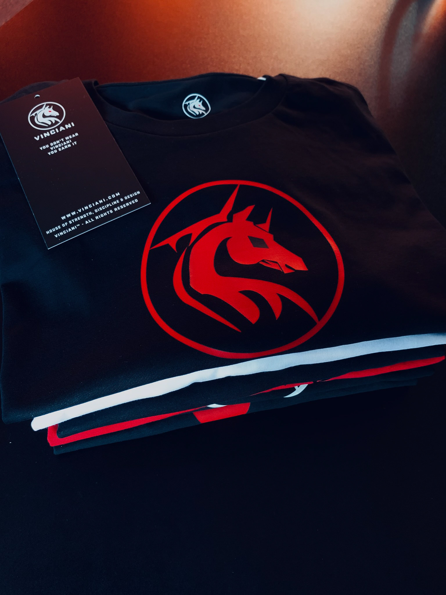 Black Vinciani luxury T-shirt folded neatly with a bold red horse emblem centered on the chest and branded swing tag attached, photographed in warm lighting.
