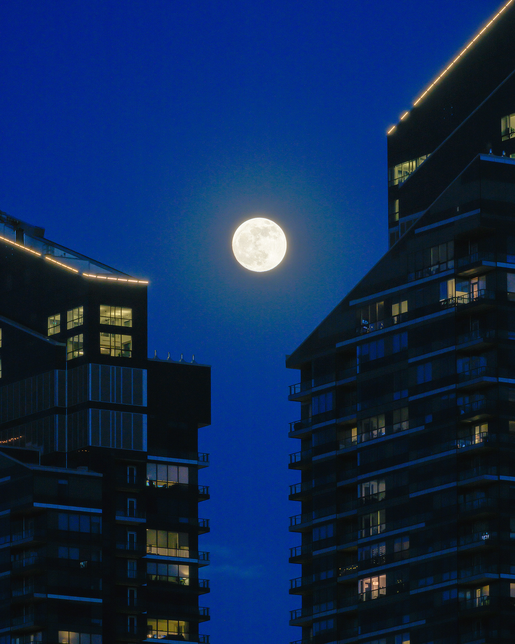 A photo of the Moon rising between a zoomed in portion of the West Village Towers during Civil Twilight, from Mewata (14 St SW) Bridge, Calgary, AB - April 22nd