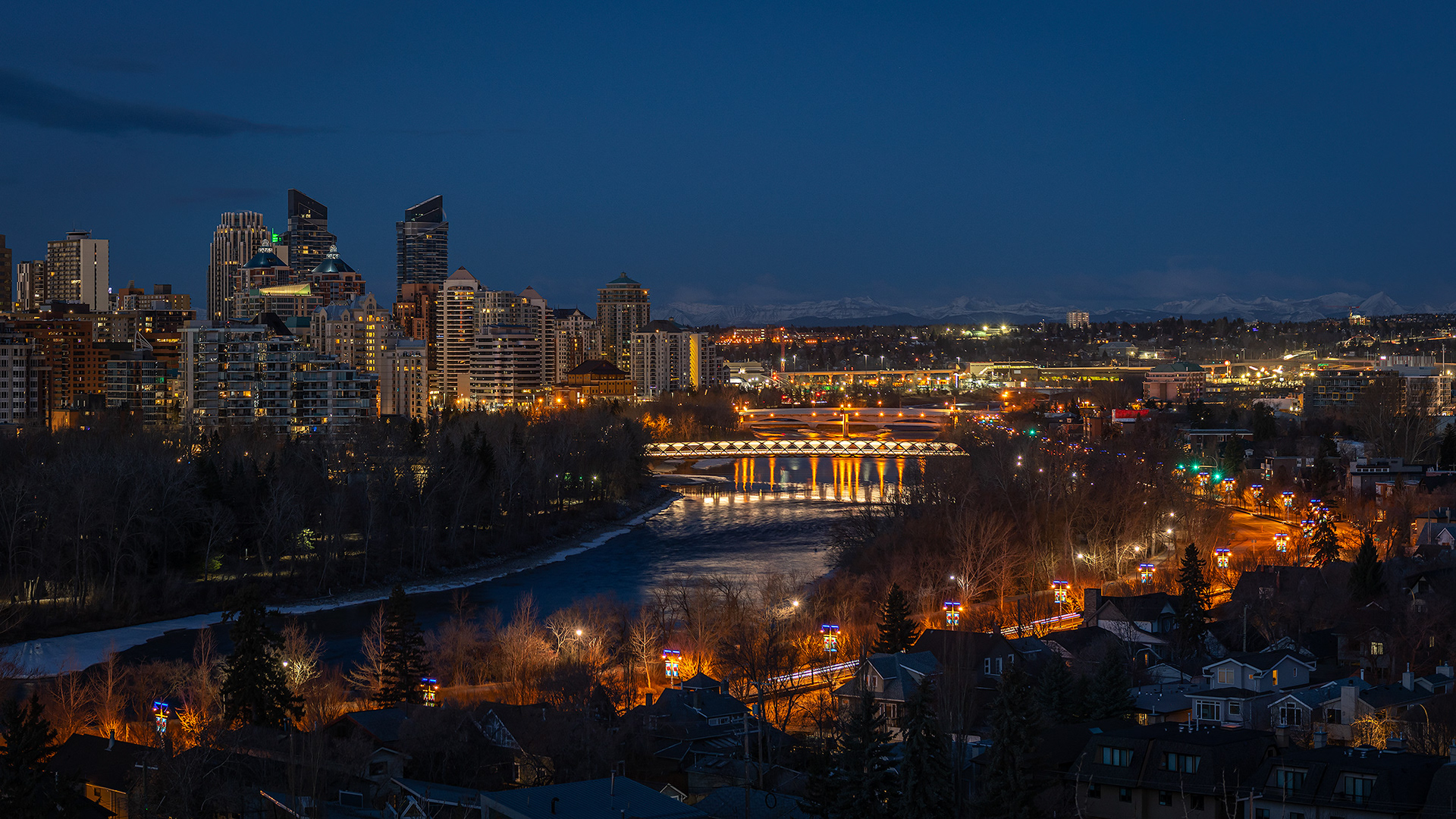 16x9 Cityscape (3 shot) of the Peace Bridge, Bow River, Memorial Dr., a portion of the downtown skyline, and the Rocky Mountains [pre sunrise, Nautical Twilight (sun altitude -7.7 degrees)], from Crescent Heights Lookout Point, Calgary, AB