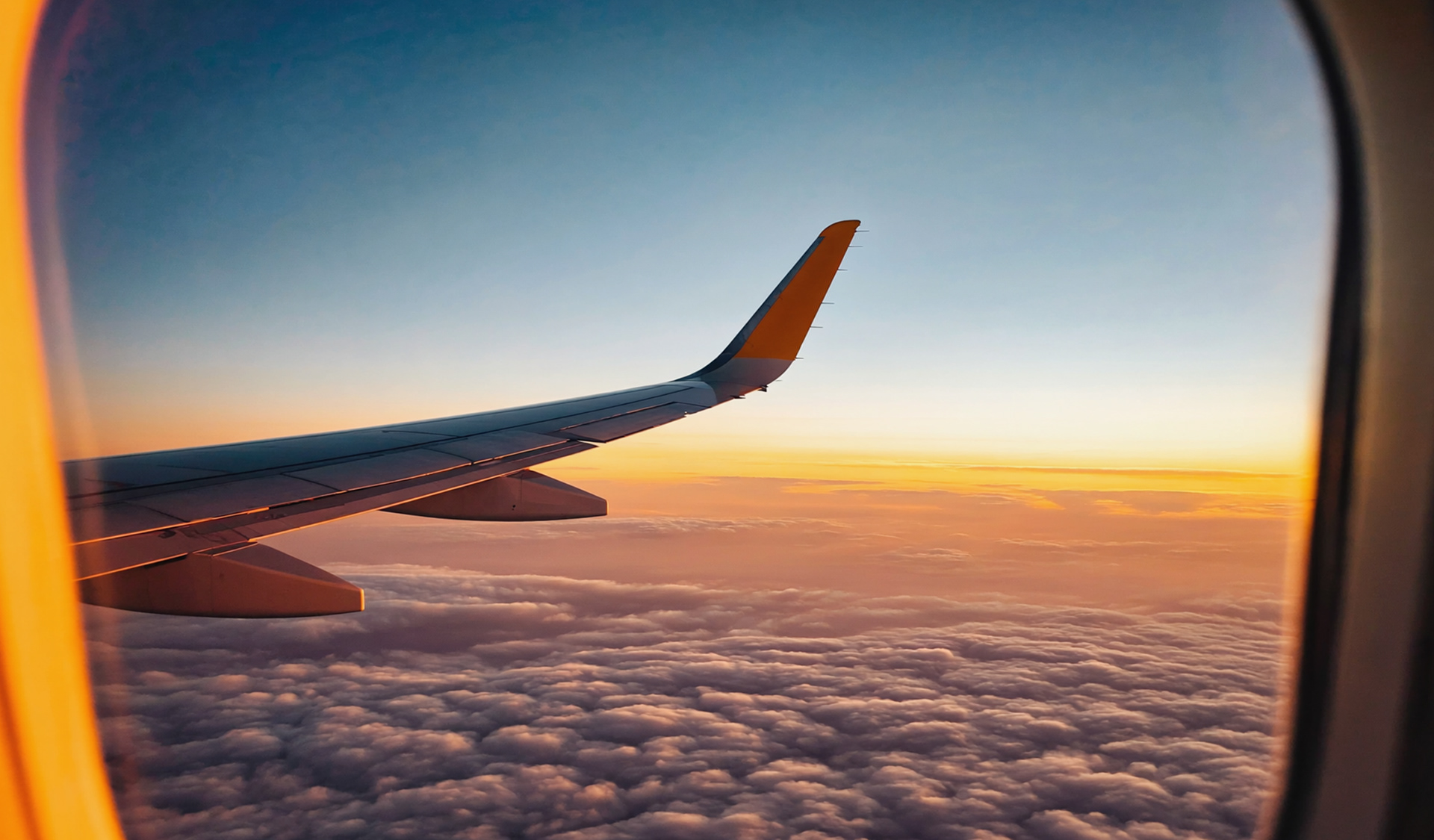 Looking out an airplane window at golden clouds and horizon line during sunset, clean aircraft wing visible, warm glow inside the cabin, minimal reflection on window, peaceful vibe, cinematic travel mood