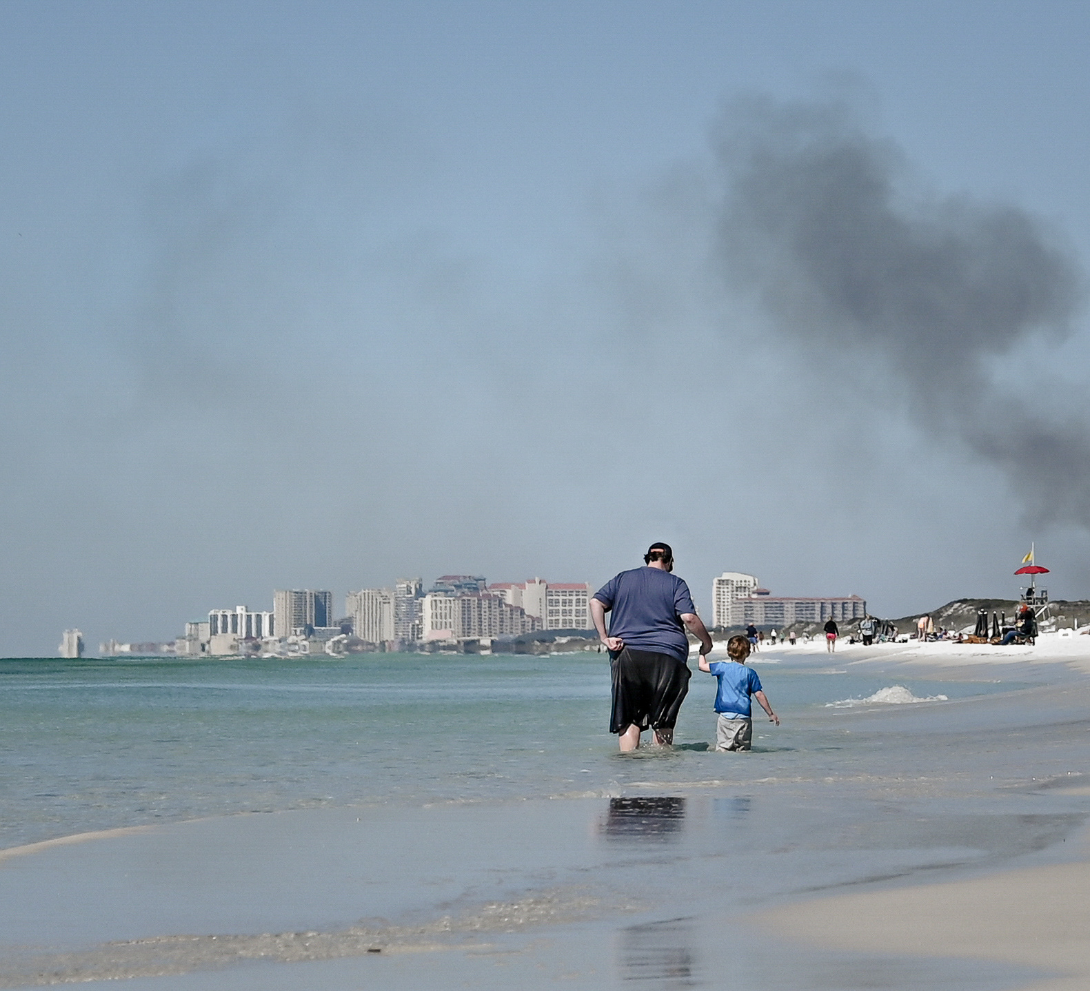 A father wades through the tide with his son in Destin, Florida while smoke from a controlled burn of Topsail Hill State Park rises in the distance.