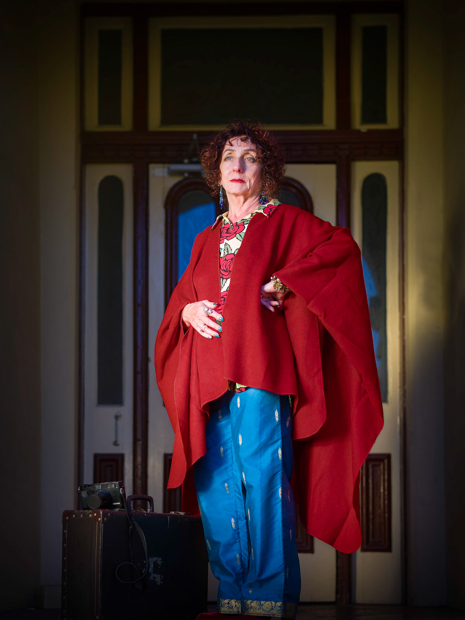 Su is a photographer, artist and traveller, she is posing at the front door of her apartment building dressed in Indian pants, a red cape over a Mambo shirt.
