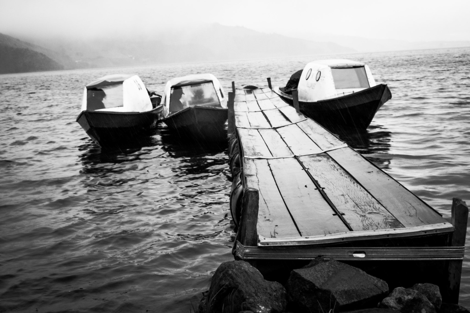 Fotografía en blanco y negro de muelle de madera con tres botes anclados sobre lago y montañas difusas al fondo