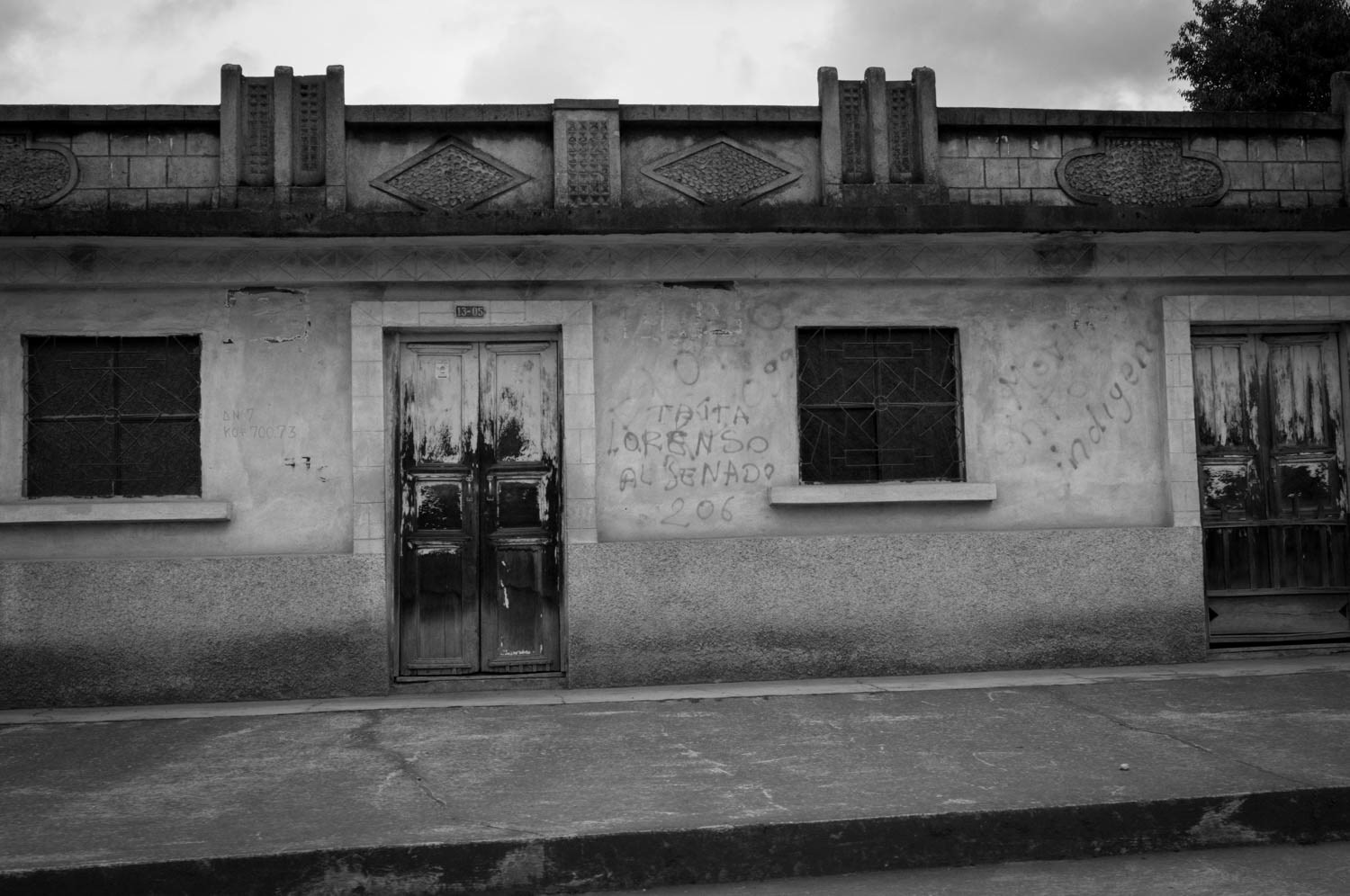 Fotografía en blanco y negro de fachada de vivienda rural con puerta de madera, ventanas enrejadas y grafitis en las paredes
