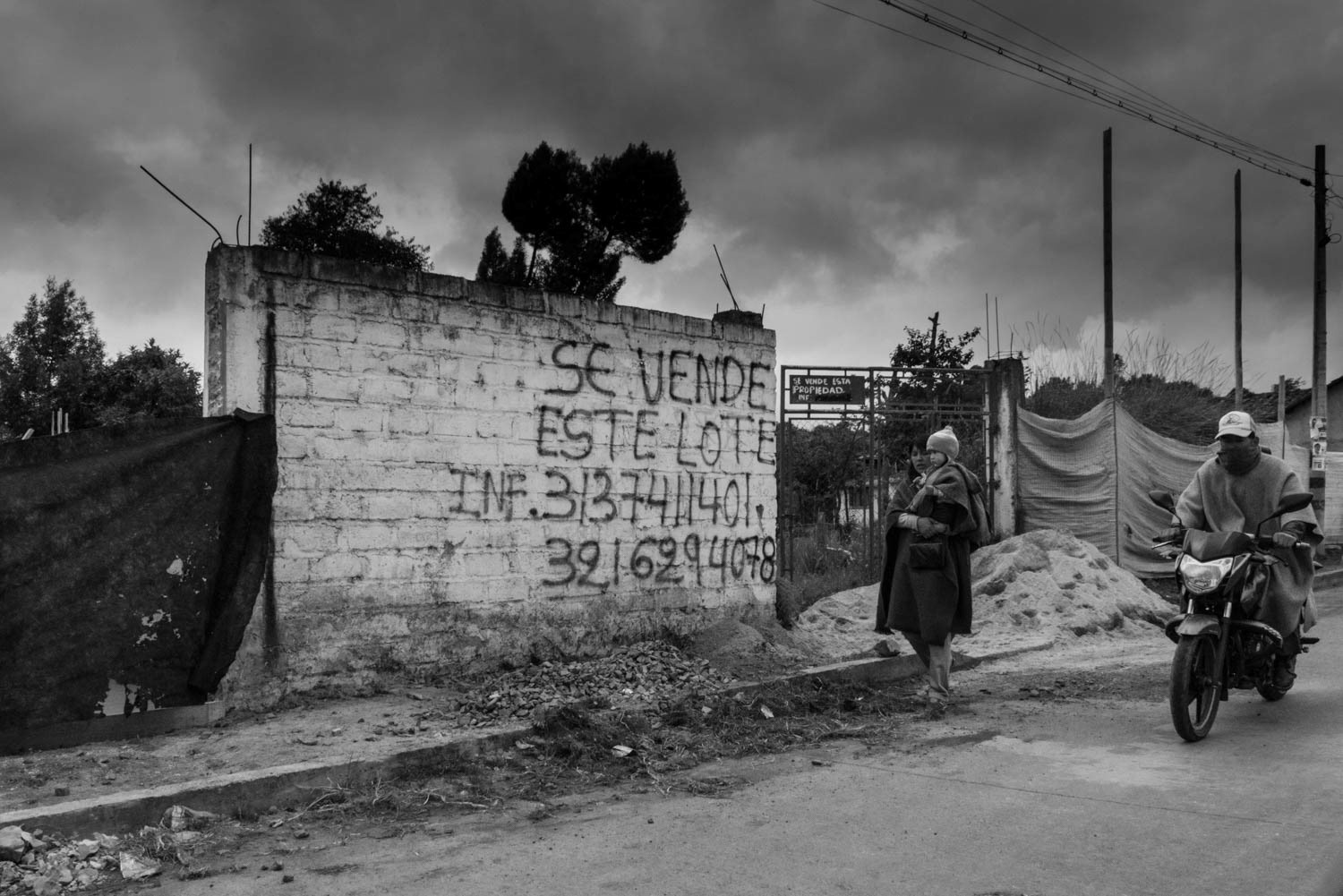 Fotografía en blanco y negro de muro con aviso “Se vende este lote”, mujer caminando y motociclista en vía rural