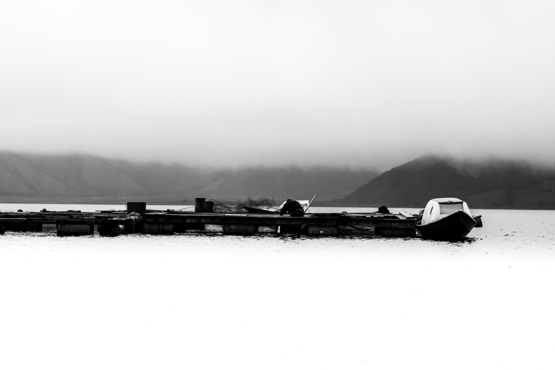 Fotografía en blanco y negro de muelle con botes sobre lago tranquilo, montañas cubiertas de niebla al fondo