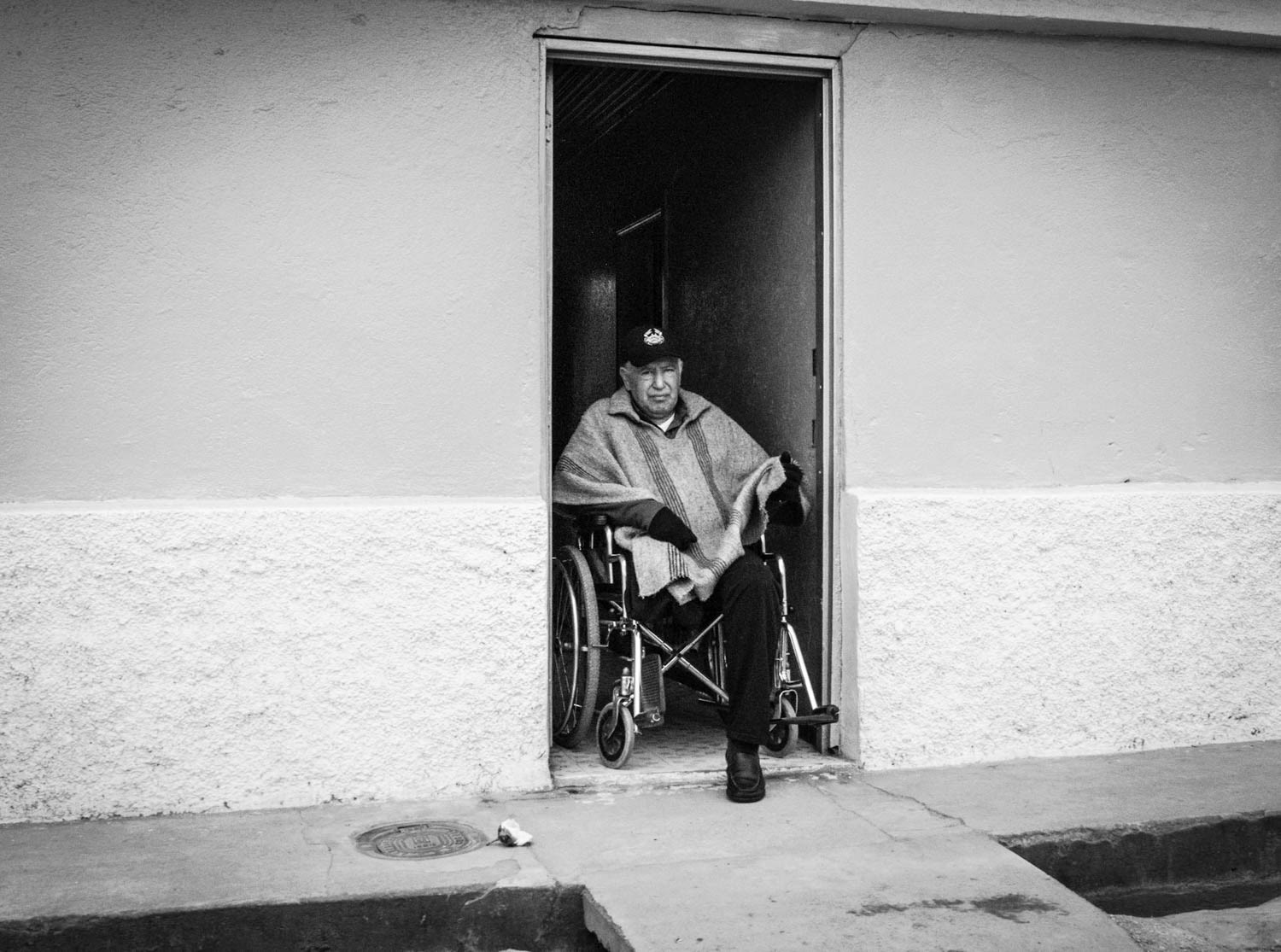 Fotografía en blanco y negro de hombre mayor en silla de ruedas sentado en el umbral de una vivienda rural