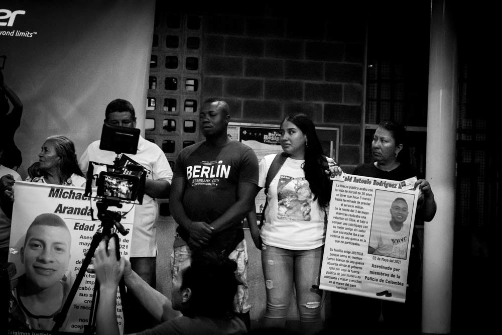 Familiares de víctimas mostrando pancartas en audiencia del Tribunal Popular en Siloé, Cali, fotografía documental en blanco y negro.