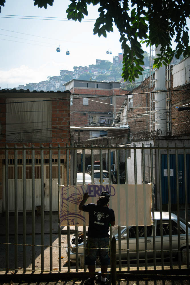 Artista urbano pintando grafiti en Siloé, Cali, con teleférico MIO Cable y viviendas en ladera al fondo, fotografía documental de arte urbano y territorio.