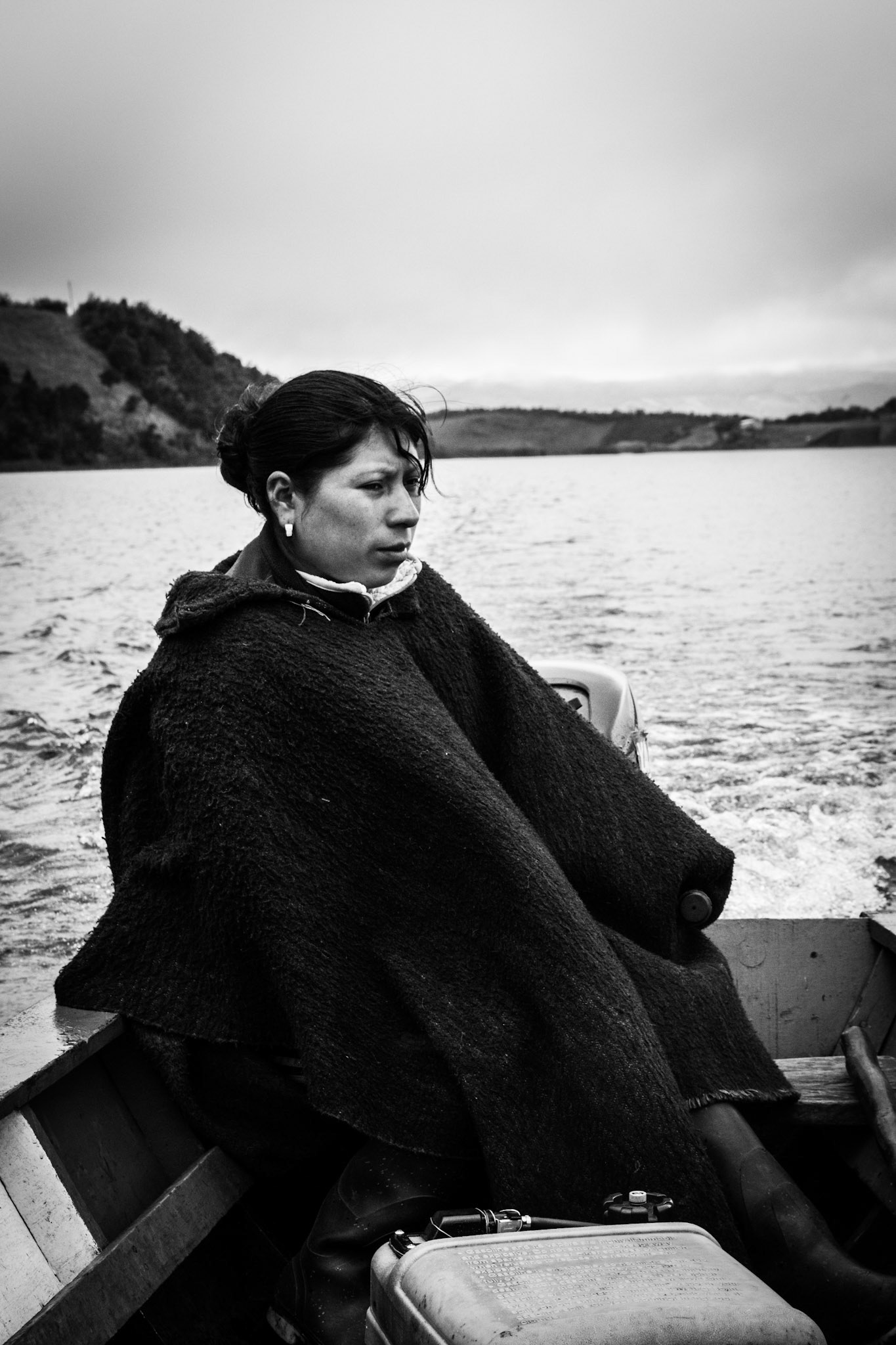 Fotografía en blanco y negro de mujer envuelta en manta navegando en bote sobre lago con paisaje montañoso al fondo