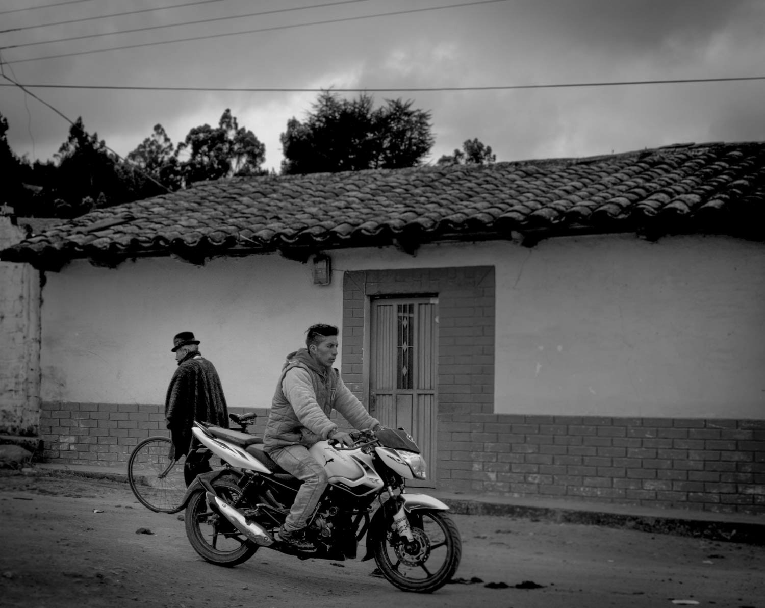 Fotografía en blanco y negro de hombre en motocicleta pasando frente a vivienda rural de adobe con techo de tejas