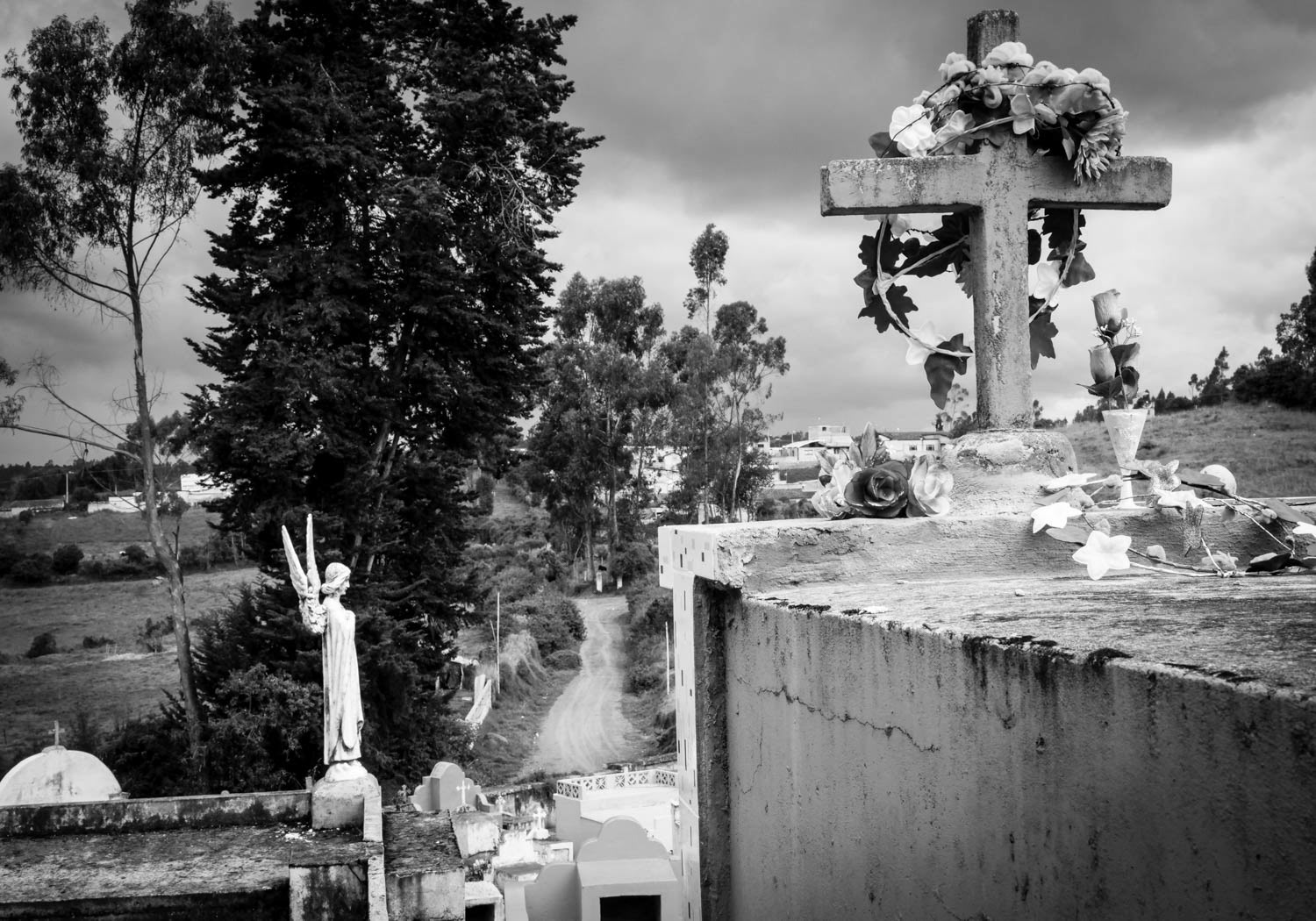 Fotografía en blanco y negro de cruces y estatuas religiosas en cementerio rural, con camino de tierra y árboles al fondo
