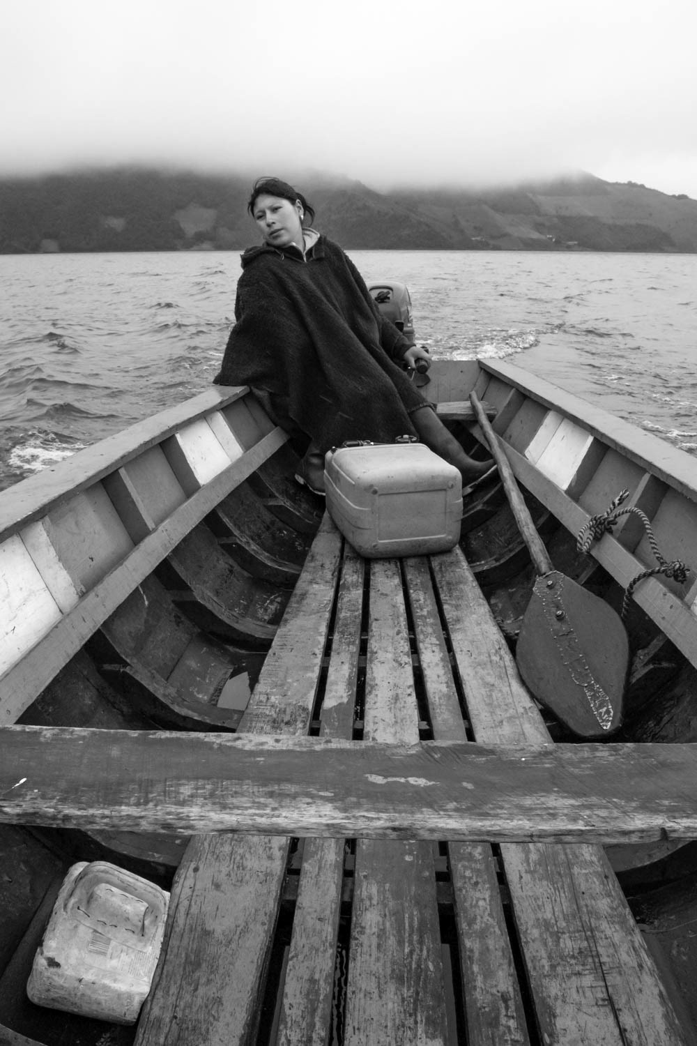 Fotografía en blanco y negro de mujer sentada en bote de madera navegando en lago con montañas cubiertas de niebla
