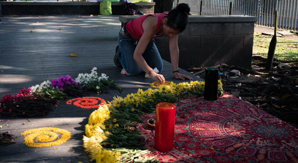 Mujer preparando ofrenda floral con velas en espacio público de Siloé, Cali, fotografía documental de ritual comunitario.