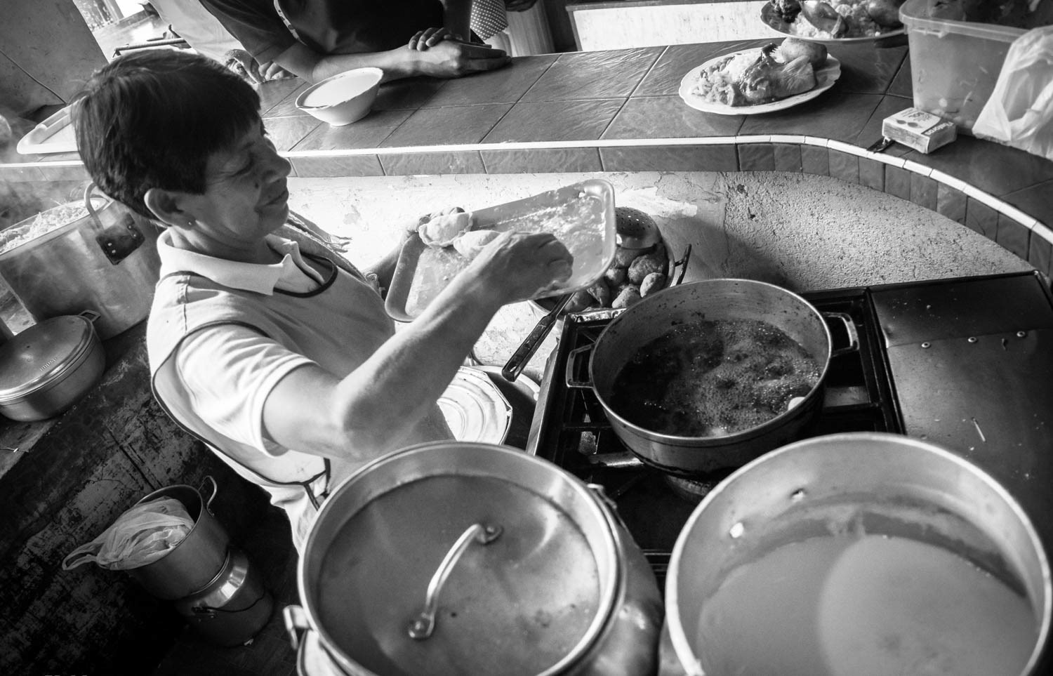 Fotografía en blanco y negro de mujer cocinando alimentos fritos en cocina tradicional, con ollas grandes y platos servidos